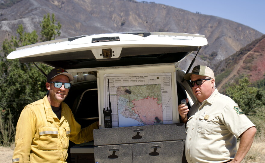 Two men stand and look at the camera in front of an open trunk. 