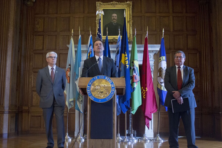 In this archive photo, Governor Steve Bullock stands at a podium with an array of state and tribal nation flags behind him. Lt. Governor Mike Cooney stands at his right hand and Montana Budget Director Tom Livers stands to his left. All men are wearing suits.