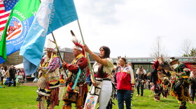 Indigenous people wearing colorful regalia hoist flags as part of the Annual Powwow of Arts and Culture.