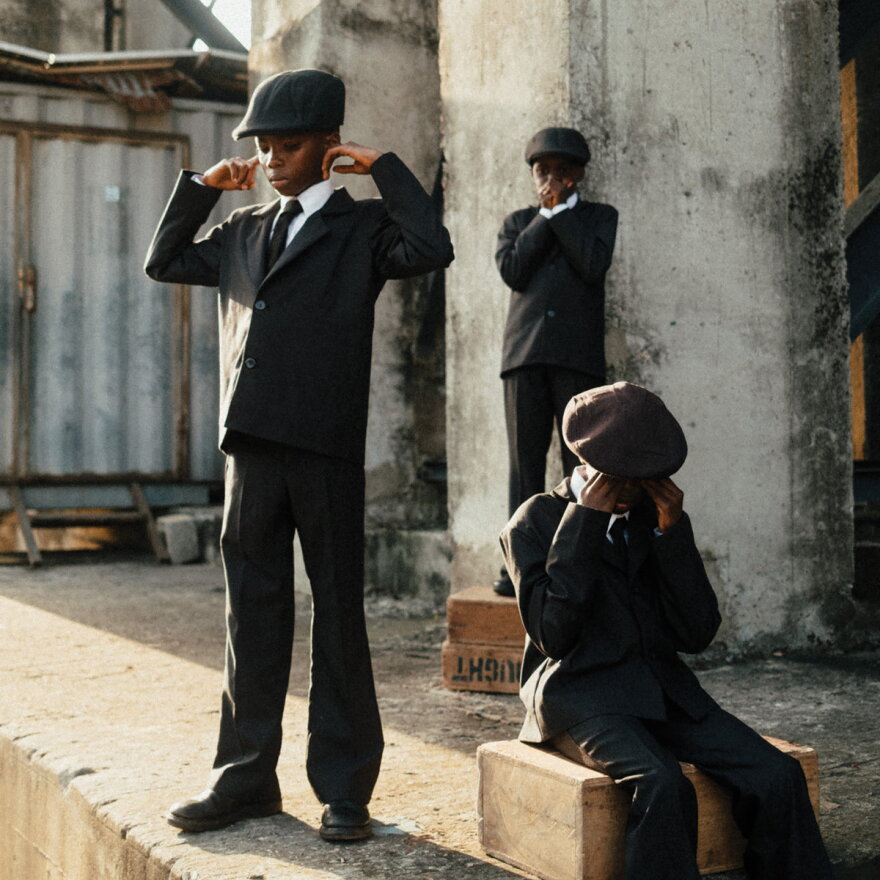 Three young boys wearing grey tweed suits with news boy caps. One is covering his mouth, one his ears and one his eyes. 