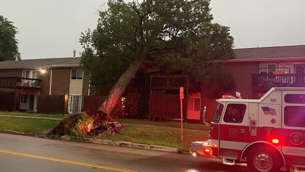 An uprooted tree falls on to a two-story apartment building with a fire truck parked on the streets with its emergency lights flashing. 