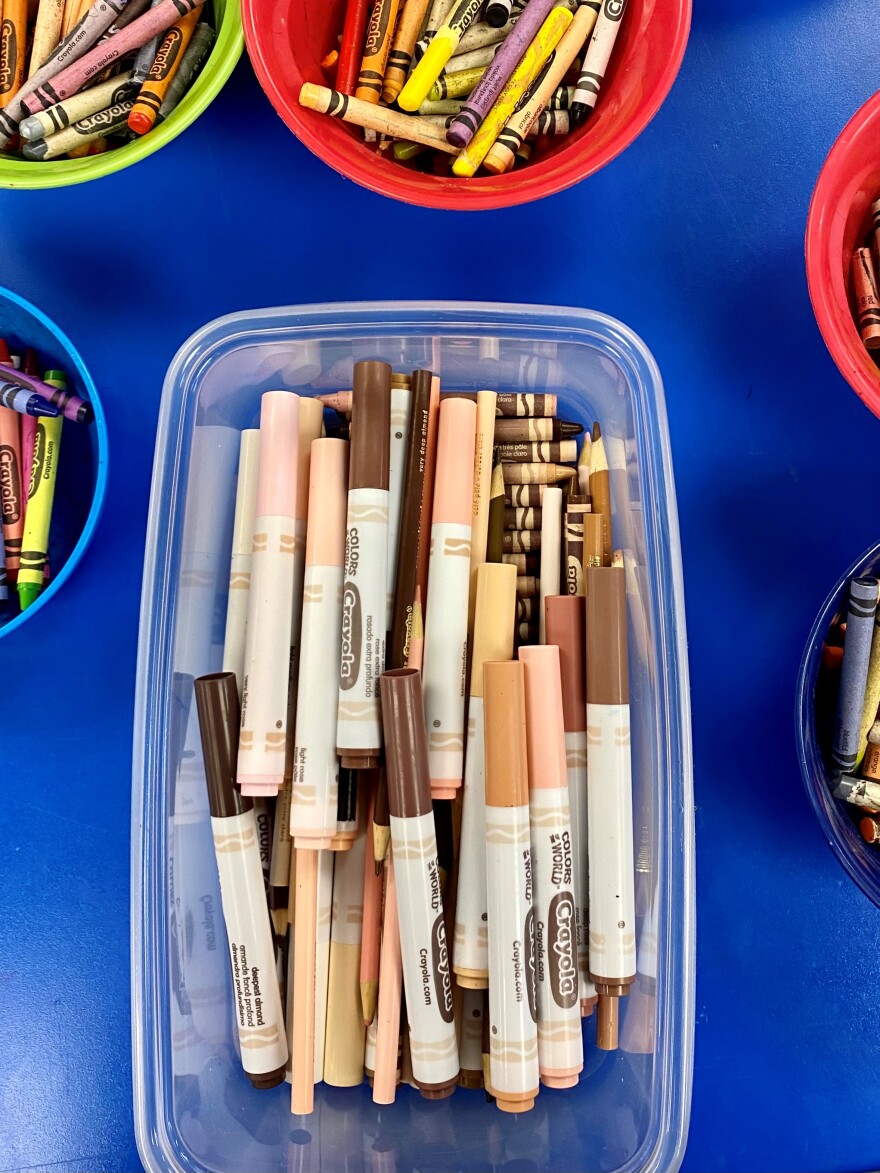 A plastic tub of crayons, colored pencils and markers in almond, gold and rose shades is surrounded by bowls of markers and crayons in primary colors.