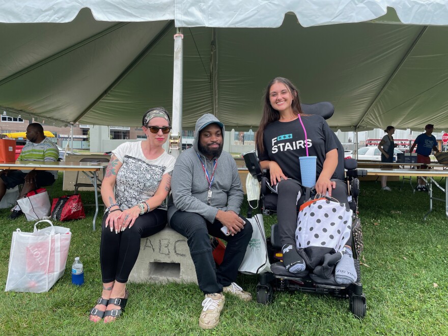 Three people pose together for a photo in front of a large event tent. One person is using a motorized wheelchair and the other two people are sitting on a concrete block.