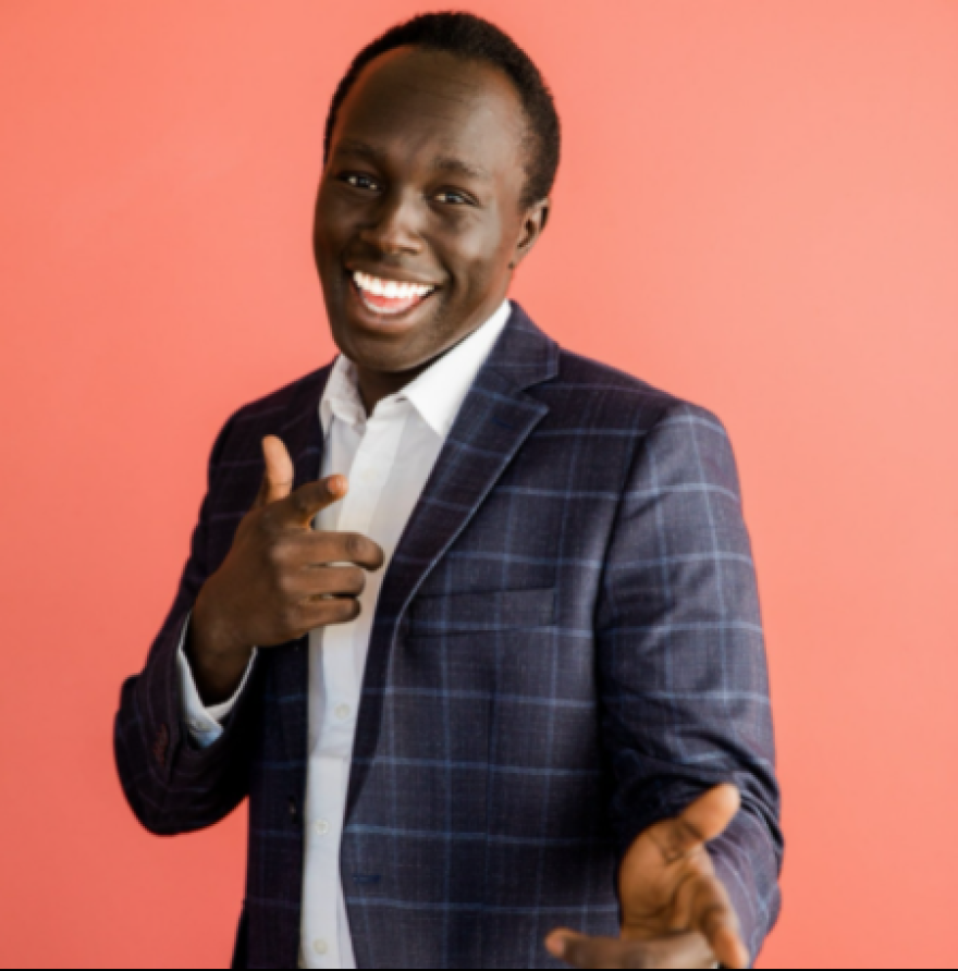  Jazari Kual Zakaria is a Black man with close-cropped dark hair. In this photo he is wearing a blue checked suit with a white shirt and is standing against a pink-orange background. He is smiling and making finger guns at the camera.