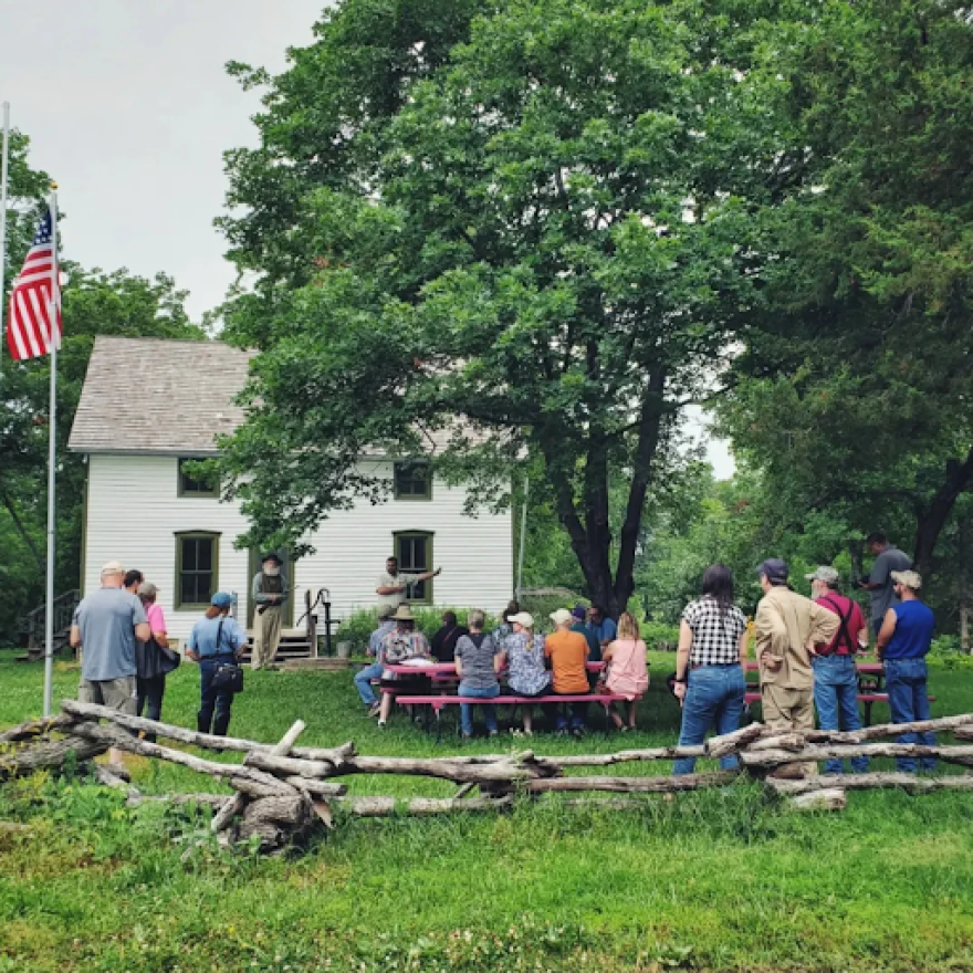 A group of people, some standing, some seated at a picnic table, listens to a man with a white beard who is standing in front of a 1800s era white wooden house.