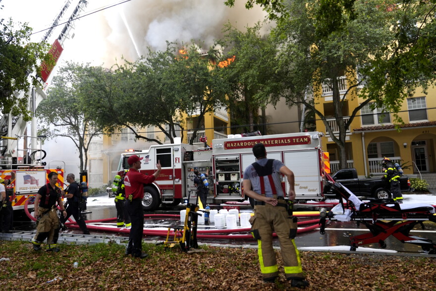 Firefighters and a fire engine in the foreground as a smoke billows out of a building.