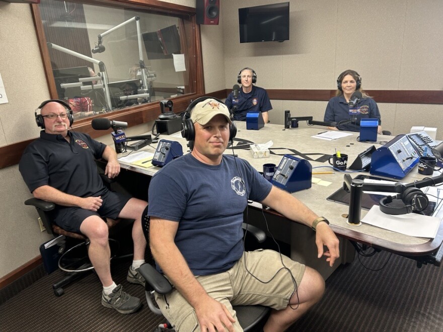 Four people wearing headphones sit around a table in a radio talk studio: A man front left is wearing a dark polo shirt with a fire department logo and shorts; a man front right is wearing a blue t-shirt with a fire department logo in white, a baseball hat, and khaki shorts; a man back left has short grey hair and is wearing a dark shirt with a fire department logo, and woman back right has brown hair and is wearing a blue shirt with a fire department logo