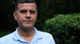An middle-aged man stares solemnly into camera, wearing a light blue polo shirt.
