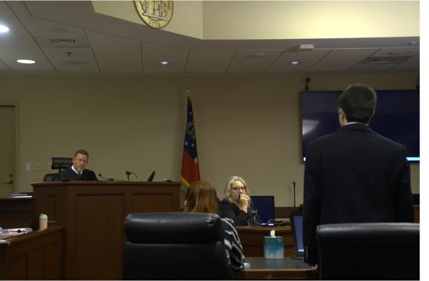 A courtroom scene shows a judge seated at the bench while an attorney stands facing him and other participants sit nearby.