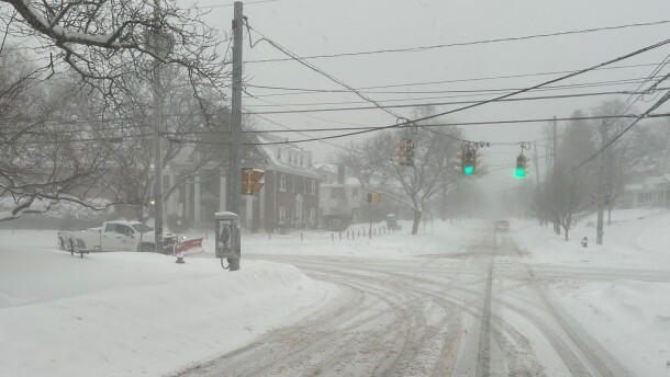 Snowy streets with a snowplow stopped at a red light, large snow banks line the streets.