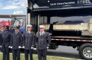 Four firefighters in navy blue uniforms stand at attention near a truck with a side panel open that shows a horizontal beam from the World Trade Center