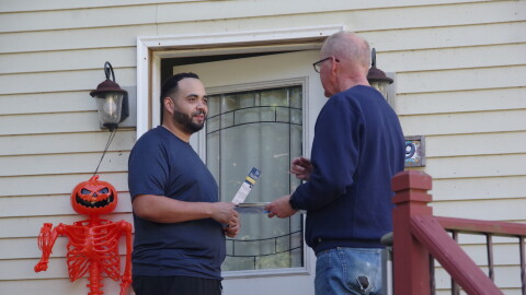 Two men talk in front of the white front door of a white wooden house. To the right of the door stands an orange skeleton with a jack-o-lantern. 