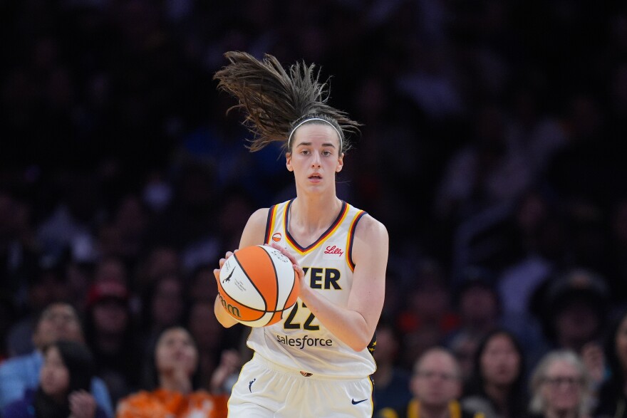 Indiana Fever guard Caitlin Clark (22) dribbles during the first half of a WNBA basketball game against the Los Angeles Sparks Wednesday, May 13, 2026, in Los Angeles. She has brown hair in a ponytail with a thin white headband. She's wearing a Fever uniform; it is white with yellow and blue stripes at the neck. 