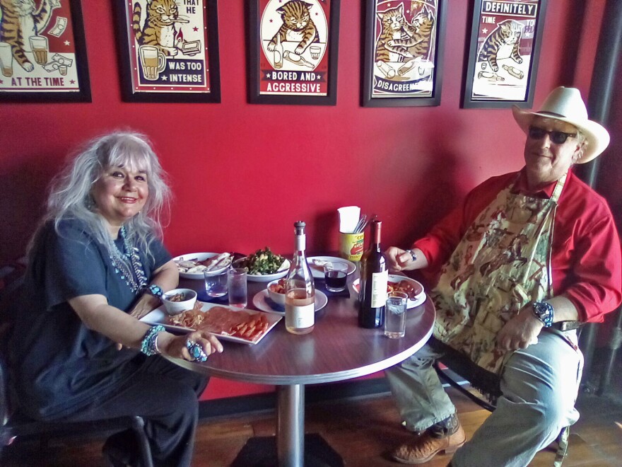 A woman with grey hair in a blue shirt and lots of turquoise jewlery sits at a table with a man in a red button down shirt, blue jeans, and a apron with horses and birds on it. They're sitting at a silver table. On the table are two wine bottles, a plate with cold cuts, a salad, some water glasses, and some other bowls. The wall behind them is bright red and has five cartoon paintings of cats drinking beer.