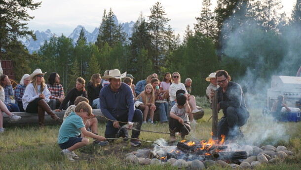 Adults and kids kneel and squat around a smoky campfire, roasting marshmallows. People sit in the background, in front of a view of rocky mountains.