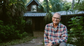 A man in a plaid long sleeved shirt is sitting outdoors on top of a decorative tiled table that's in front of a forrested cabin
