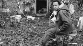 A man sits in a beat-up metal chair in a forest clearing and looks at the camera with a damaged shed in the background. 