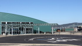 An airport terminal building overlooks a bay with a cantilever bridge in the background and a painted parking lot in the foreground with "Fly OTH" pained on it.