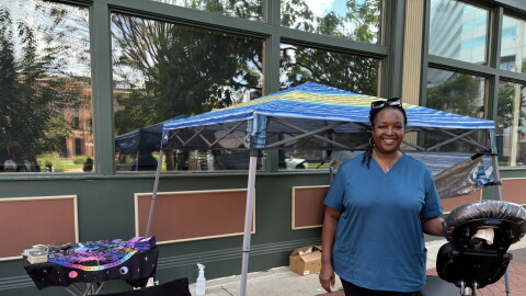 Woman in blue top and dark pants smiles for a portrait while standing under a pop-up awning, next to a massage chair