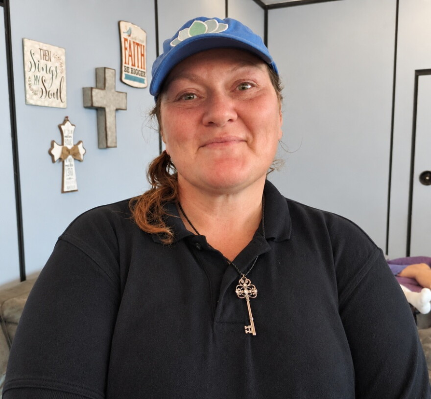 caucasian woman in blue baseball cap with shoulder length pony tail coming out the real, wearing black polo shirt  with an old-fashioned key on a necklace around her neck . behind her is a light blue wall with two artsy crosses and religious-saying art hanging on the wall 