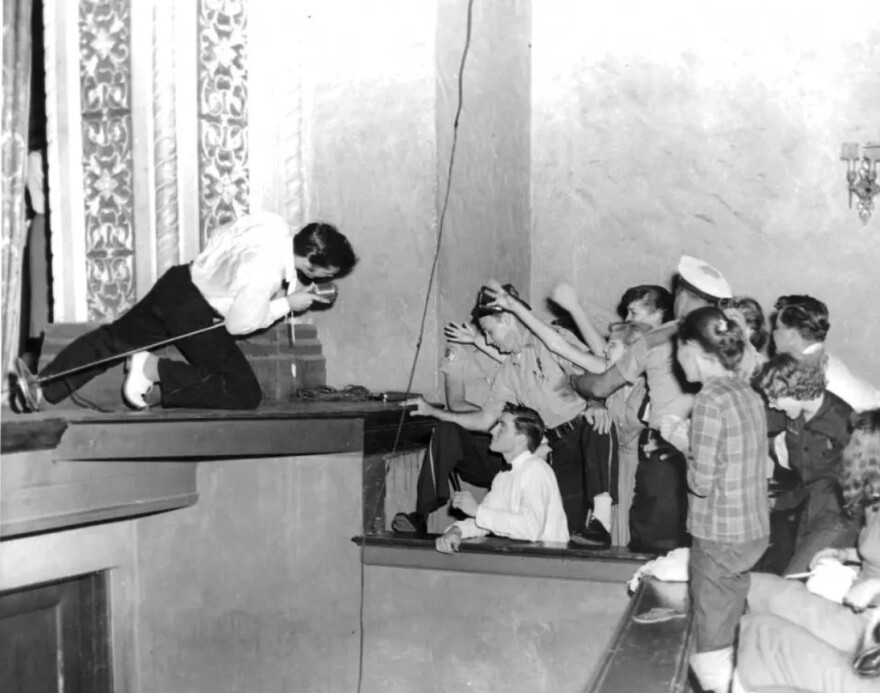 Elvis sings into a microphone on his knees onstage in front of a crowd. 
