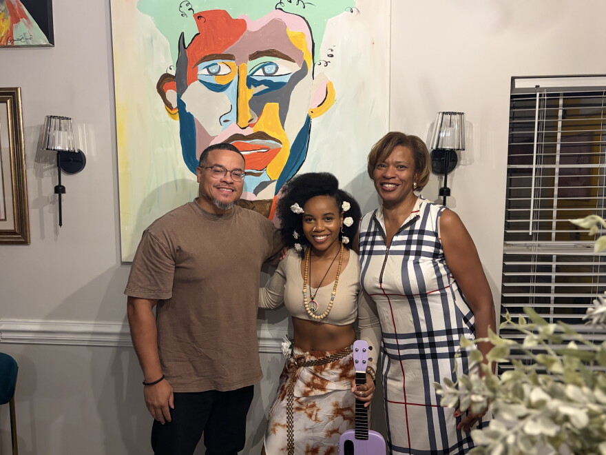 One Black man and two Black women smiling in room in front of wall painting