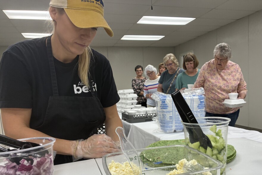 In the foreground, a woman prepares food while seniors walk in line to grab food in the background.