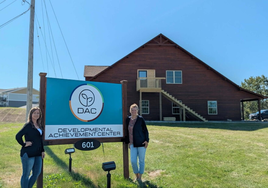 Two women stand on each side of a blue sign that reads "Developmental Achievement Center" in front of a wood sided building.