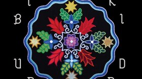A black square with an Anishinaabe multi-colored floral design. There is a wavy blue circle around the design. Over the art is written "Bizhiki" and under it is written "Unbound."