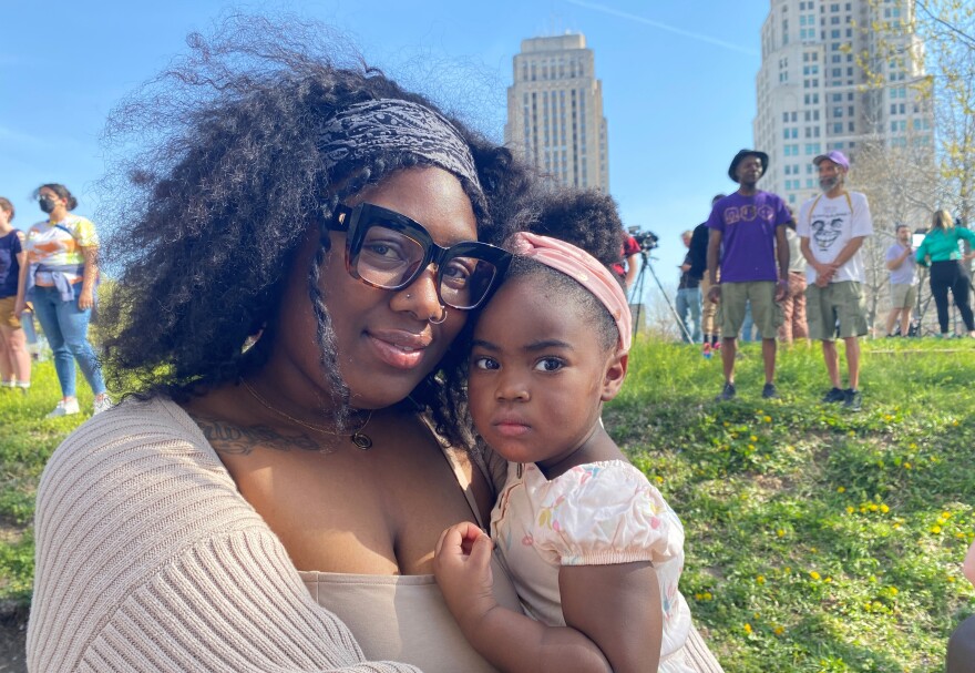 A Black woman wearing a beige sweater, glasses and a head scarf holds her young daughter, who is wearing a peach dress and headband. In the background is the Kansas City, Missouri, city hall building