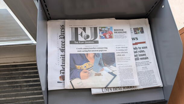 The Flint Journal sits on top of a copy of The Detroit News and the Detroit Free Press in a grey, metal newsstand