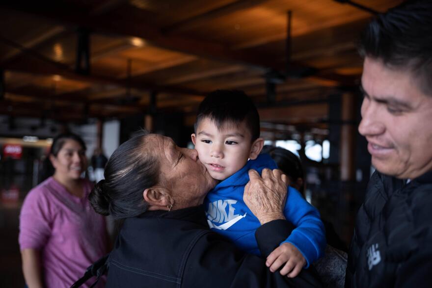 A woman holds a toddler and kisses his cheek. A  few other people stand nearby.