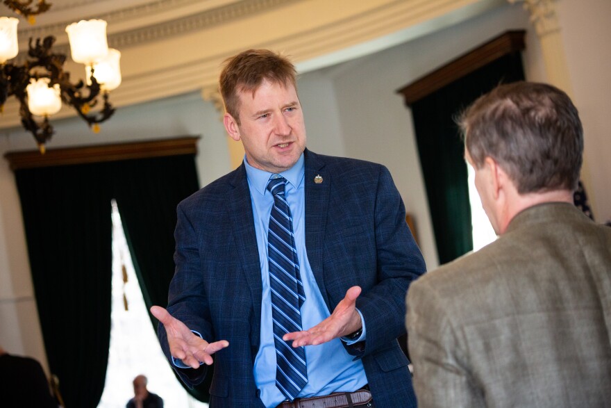 A white man with light hair and a dark colored suit speaks to someone else and gestures with his hands.