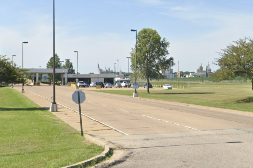 The entrance to the chemical plant has a checkpoint for employees and a parking lot — presumably for visitors. Towering industrial buildings are in the background. 