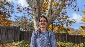 A woman with a stethoscope around her neck smiles under a tree.