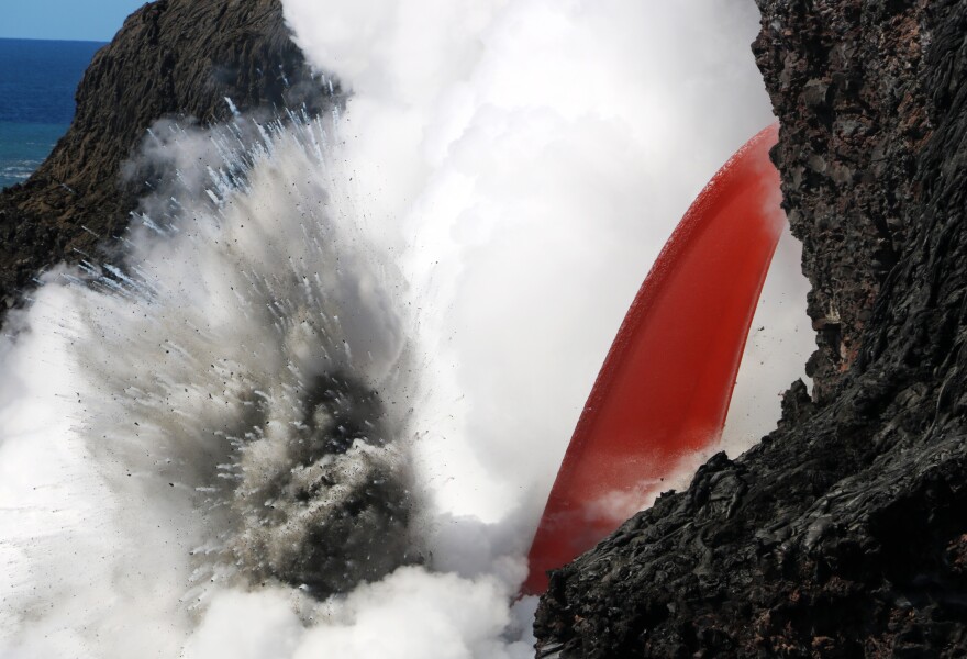 An open lava stream pours out of the lava tube, perched high on the sea cliff, and into the ocean.