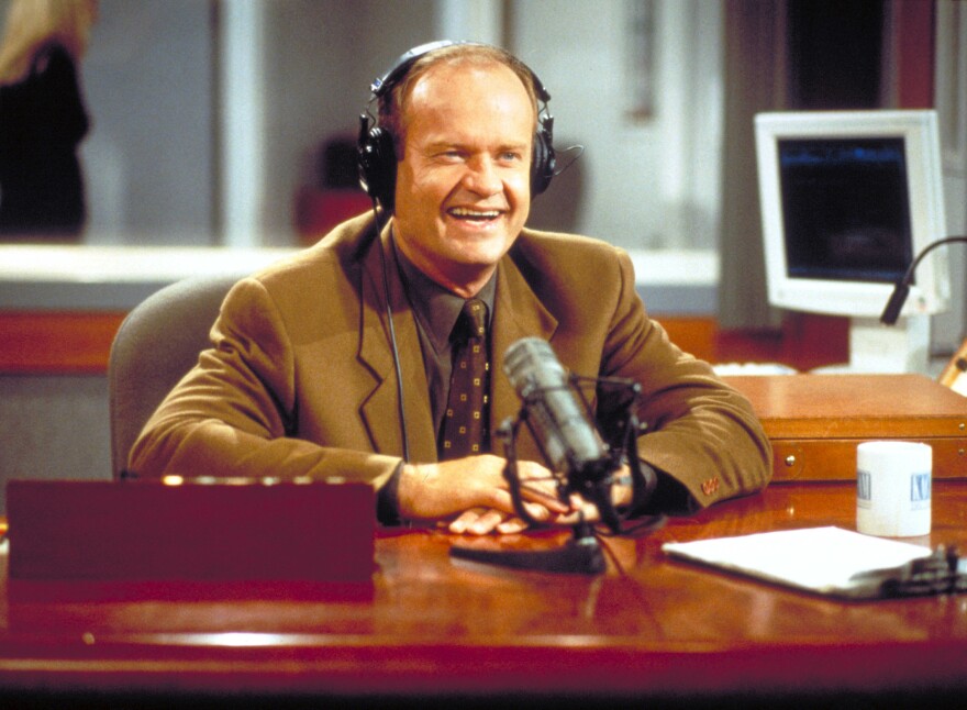 A smiling man in headphones sits at a desk with a microphone.