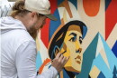 A close shot of the artist, a bearded white man, leaning towards his mural, painting the details of a face looking in the distance, and wearing a helmet. 