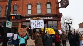 A small group of people stand in front of a crosswalk in downtown Saline. One woman in the center of the image holds a sign that reads “No Secret Deals.”