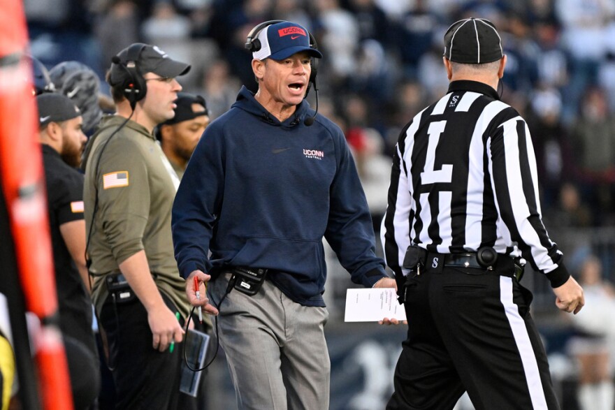 A man in a blue hoodie walks on the sideline of a football field.