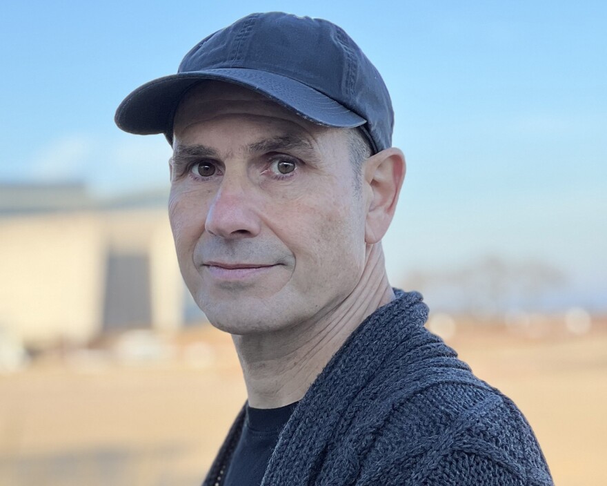 A professional headshot of author Naeem Murr, who is wearing a navy cardigan, black t-shirt and navy baseball cap. His body is angled to the left side of the image, with his face turned toward the camera.