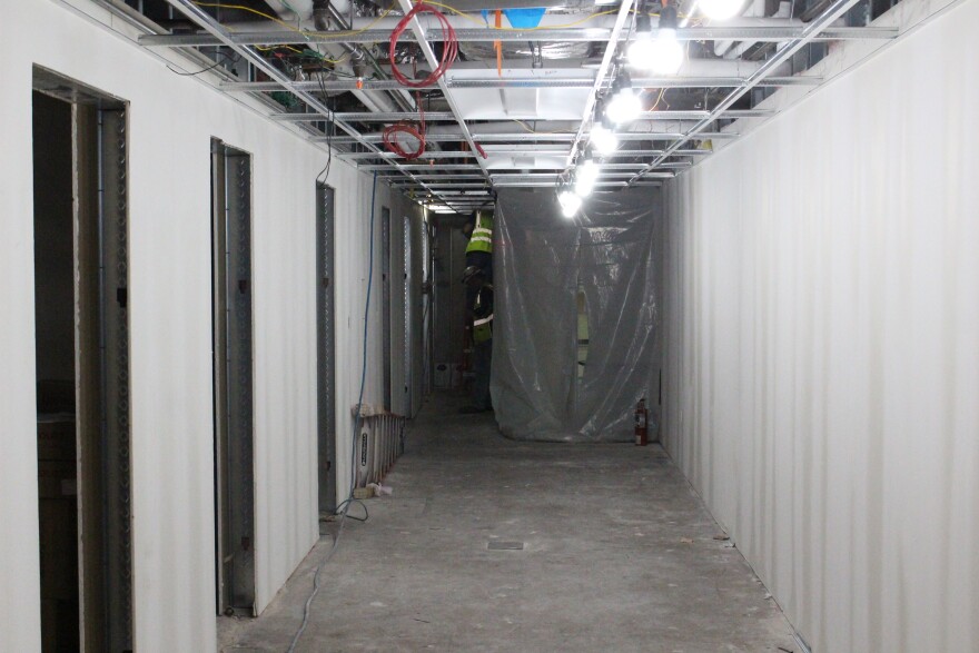 A hallway lined by drywall, showing exposed ceiling. Plastic drapes and workers are in the distance. It's a construction site.