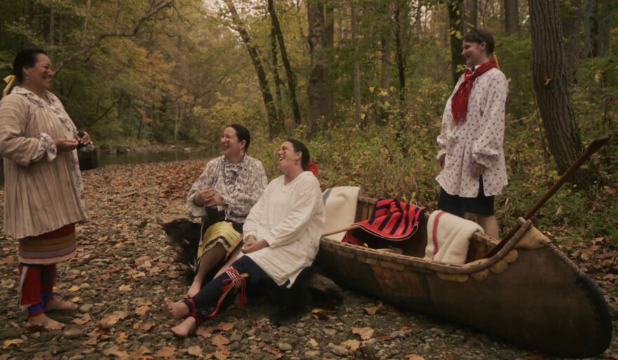 Four individuals smile and laugh while in a forest near a body of water. Two of the individuals are leaning against a boat, while the other two are standing. 