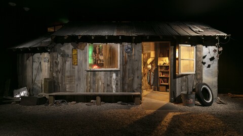 A small ramshackle cabin made out of wood sits in an exhibit in an art museum. The cabin is well-lit, but the surrounding space is made to look like night.