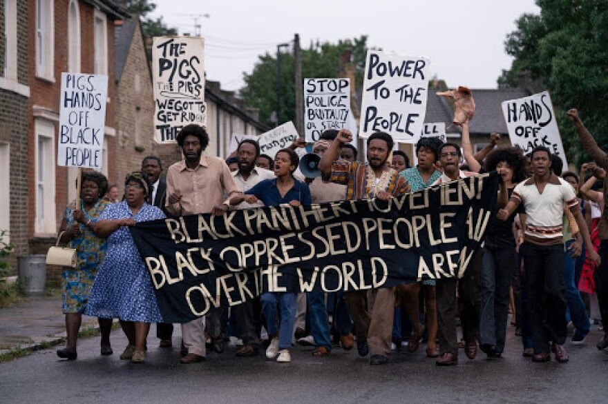 A group of Black people in the middle of the street, walking. The people are holding various signs and there is one large black banner with the words 'Black Panther Movement Black Opressed People All Over The World' in bold, gold letters