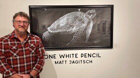A man with curly salt and pepper hair and glasses clasps his hands across his red, plaid shirt. Behind him is a black and white drawing of a sea turtle and script on a white wall in black block lettering. The text reads: One White Pencil. Matt Jagitsch