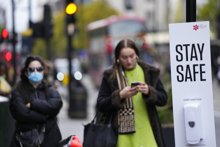 Bus passengers wait at a bus stop next to a Stay Safe sign which encourages social distancing and the wearing of masks to curb the spread of COVID-19, in London, Tuesday, Nov. 30, 2021.