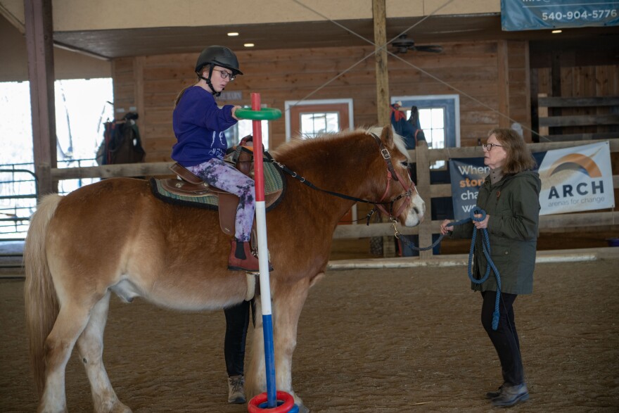 Maci Winebarger sit on a brown horse. She holds a green ring in her hand which is attached to a pole, as an instructor looks on. They're inside a large barn at Healing Strides, an equine therapy facility in Franklin County, Virginia. 