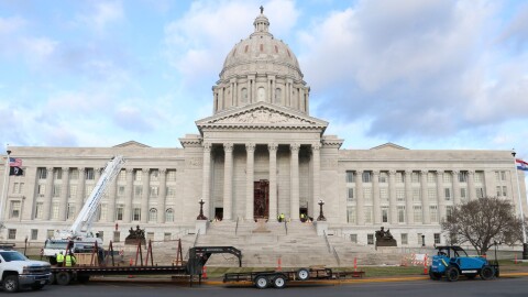 Some trucks and trailers are parked outside the Missouri capitol building. The sky is blue with white clouds. 