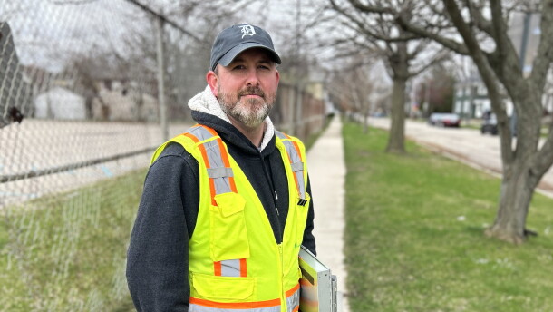 Man in a ballcap and greenish-yellow safety vest with clipboard under one arm smiles subtly while facing the camera